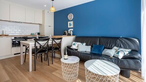 A modern kitchen with white cabinets, a dining area with a white table and chairs, a black leather sofa with patterned cushions, and a round white coffee table.
