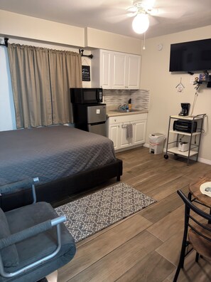 Een kleine slaapkamer met een bed, een kitchenette, een bureau en een stoel.