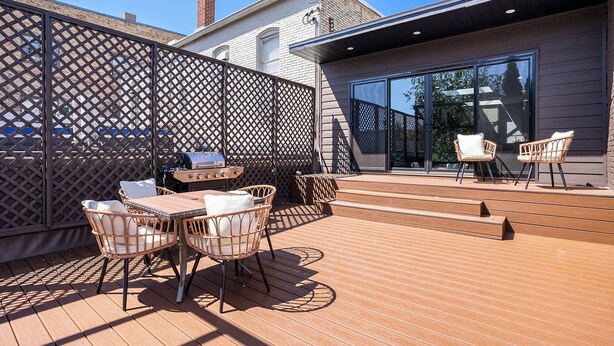 Un patio exterior con terraza de madera, sillas de mimbre, una mesa y una parrilla.