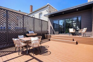 Un patio exterior con terraza de madera, sillas de mimbre, una mesa y una parrilla.