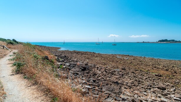 Ein felsiger Strand mit einem deutlichen Pfad zum Wasser, Boote in der Ferne und einem strahlend blauen Himmel.