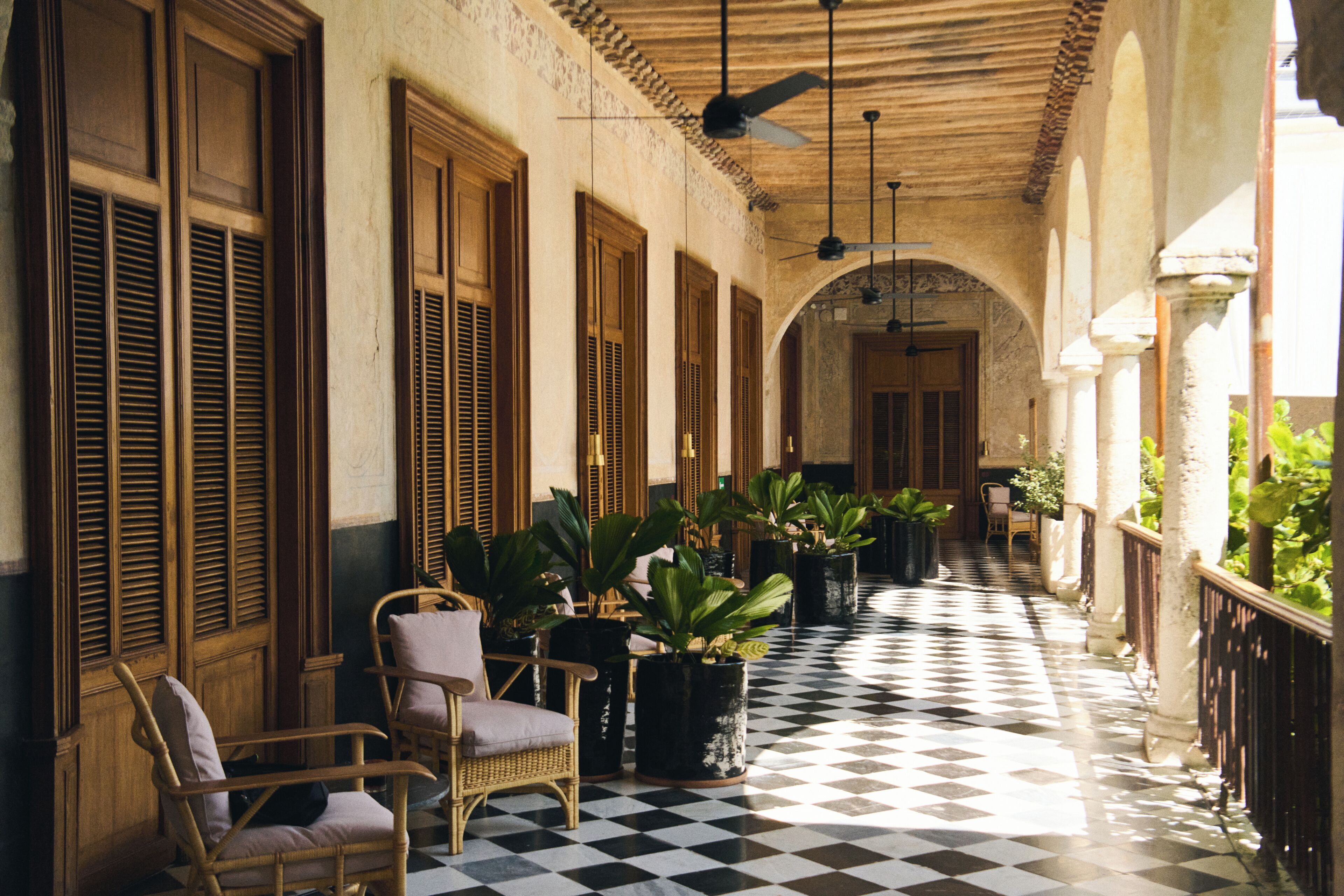 A corridor with checkered flooring, wooden shutters, and potted plants.