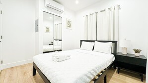 A modern bedroom with a bed, a bedside table with a lamp, a mirror, and an air conditioning unit.