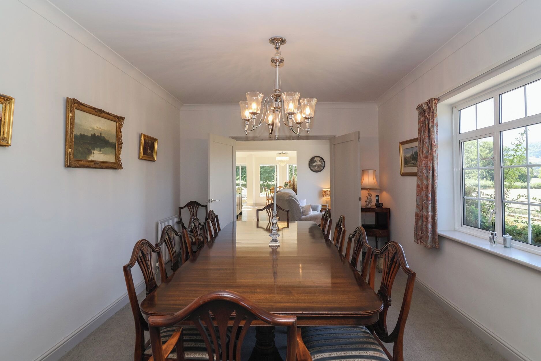 A dining room with a long wooden table, chairs, and a chandelier. There are framed pictures on the walls and a window with curtains.