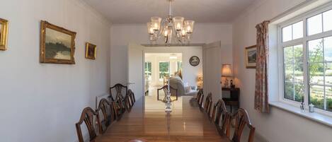 A dining room with a long wooden table, chairs, and a chandelier. There are framed pictures on the walls and a window with curtains.