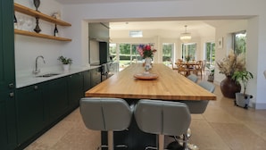 A modern kitchen with a central island, dark green cabinets, and a dining area with natural light. - Leafield Cottage (Gloucester)
