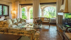 A cozy living room with a floral sofa, a coffee table, and a view through an arched doorway to a garden. - Dolce Collina 10, Emma Villas (Cesena)