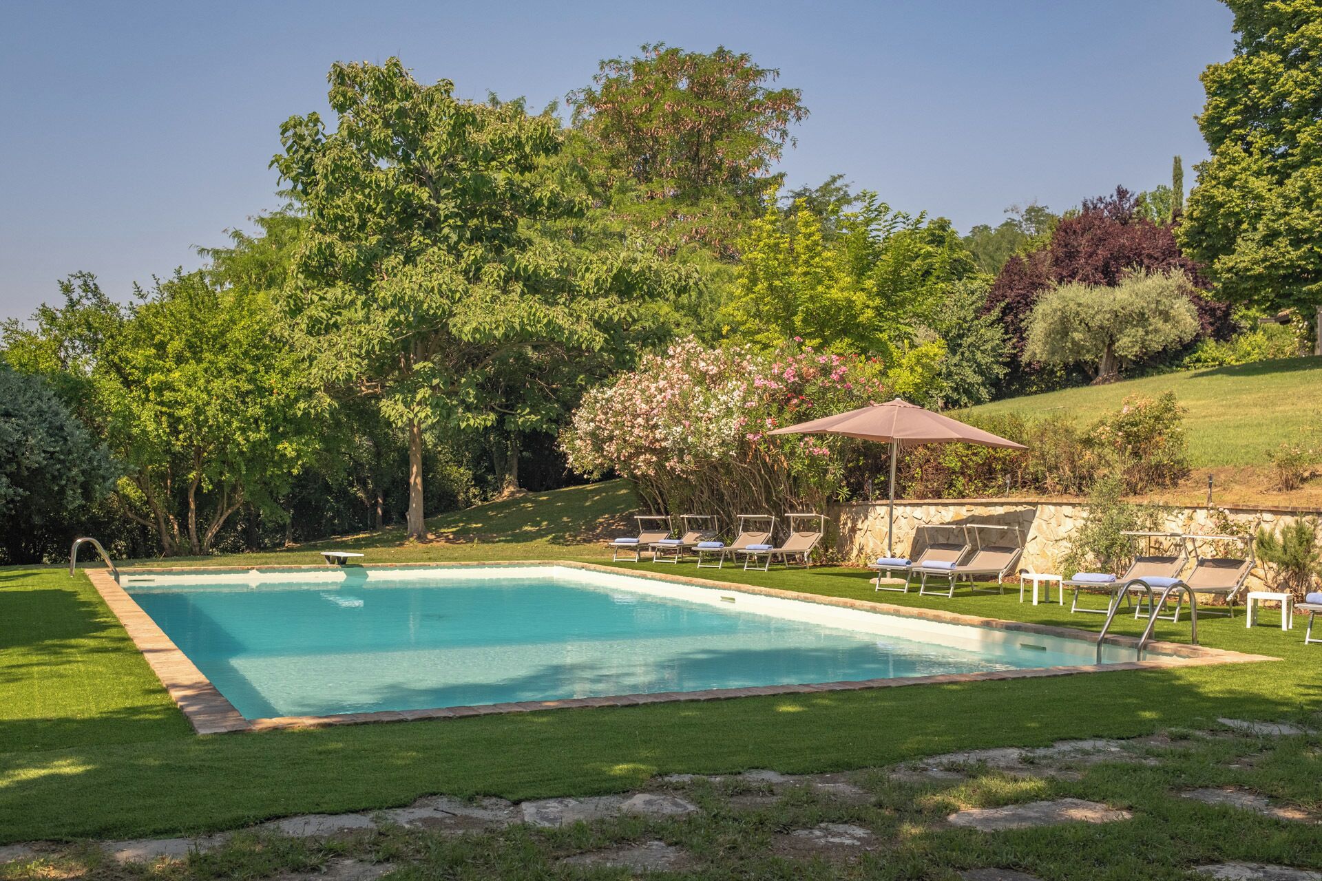 A rectangular swimming pool with a stone border, surrounded by well-maintained grass. There are lounge chairs with umbrellas and a stone wall in the background.