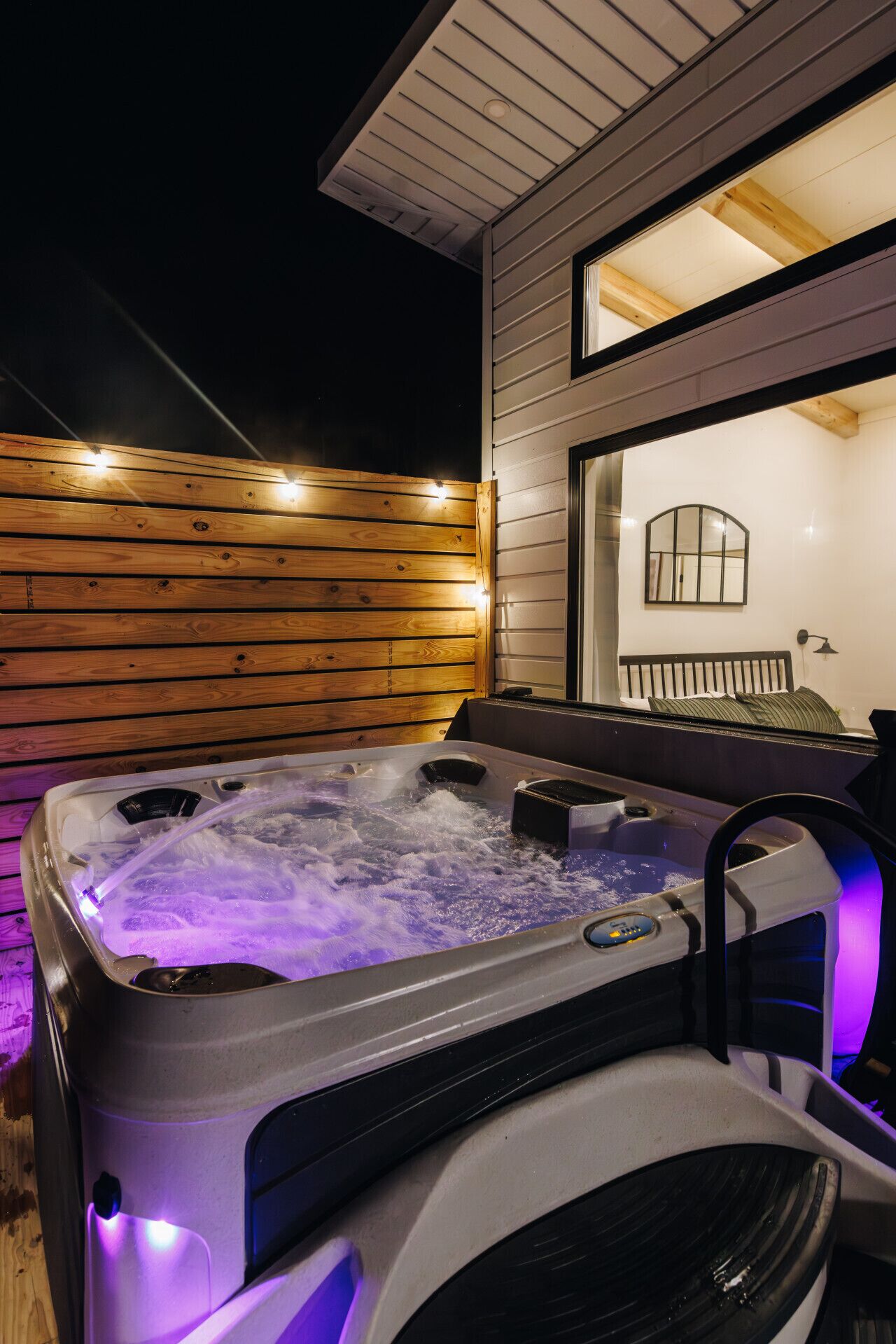 A hot tub with a control panel, surrounded by wooden decking and a white wall with a window.
