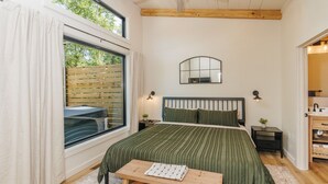 A bedroom with a large bed, a wooden bench, a nightstand, a mirror, and a window with curtains. - Modern Silo | Hot Tub | Trailer Friendly | Dogs (Beattyville)
