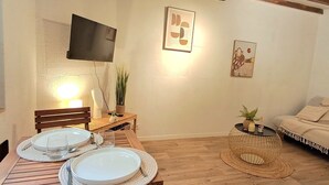 Un coin repas avec une table en bois dressée pour deux personnes, un téléviseur fixé au mur et un canapé en arrière-plan.