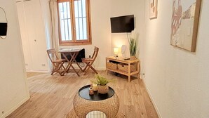 Une pièce chaleureuse avec une table à manger et des chaises en bois, une petite table d’appoint avec une lampe et des objets décoratifs, et un téléviseur fixé au mur.