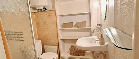 A small bathroom with a toilet, a sink, and a mirror. There are shelves above the sink and a basket on the floor.