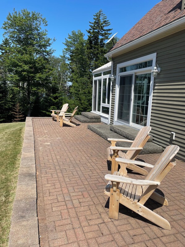 En terrasse med træ-Adirondack-stole og en murstenssti, der fører hen til et hus med en overdækket veranda og store vinduer.