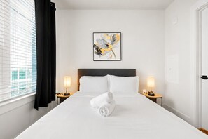 Ein Hotelzimmer mit einem großen Bett, zwei Nachttischen mit Lampen, einem gerahmten abstrakten Gemälde und einem Fenster mit Vorhängen.