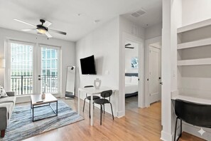 Uma sala de estar moderna com sofá, mesa de centro e televisão. Há uma varanda com vista, uma área de jantar com cadeiras e um quarto visível por uma porta aberta.