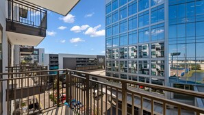 A modern building with a glass facade, a balcony with metal railings, and a parking area with cars.