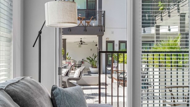 A modern living room with a grey sofa, a hanging light fixture, and a view of an outdoor balcony with a dining area and plants.