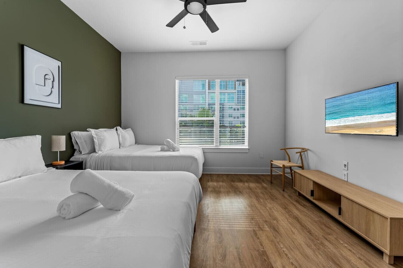 Et moderne hotelværelse med to senge, trægulv, stort vindue med persienner, ventilator i loftet, tv på væggen og en træbænk.
