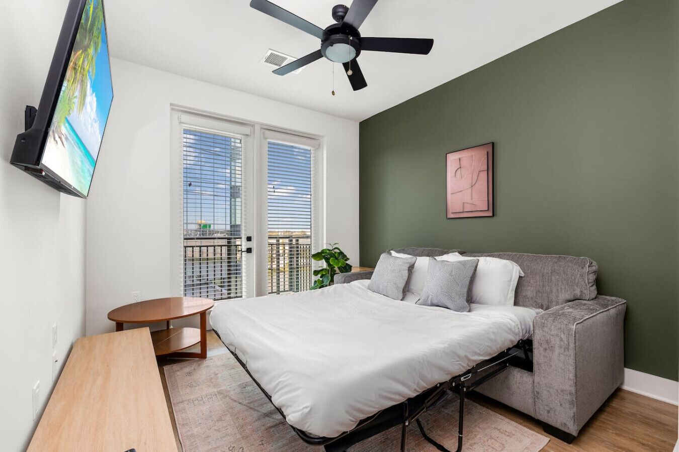 Ein Schlafzimmer mit einem Bett, einem grauen Sofa, einem kleinen Tisch, einem Deckenventilator und einem an der Wand befestigten Fernseher.
