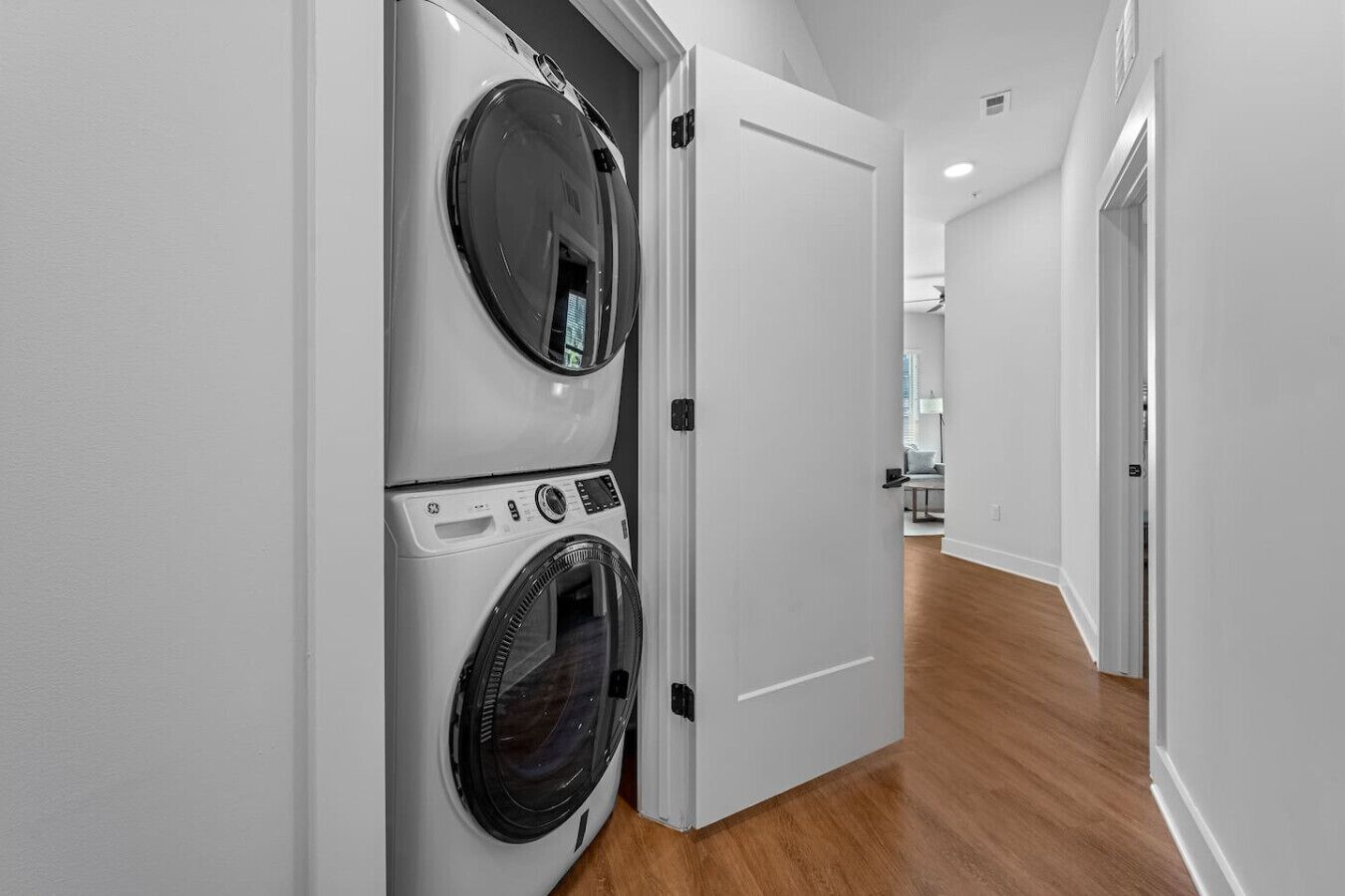 Un cuarto de lavandería con dos lavadoras de carga frontal apiladas y una puerta que da a un pasillo con piso de madera y una lámpara en el techo.