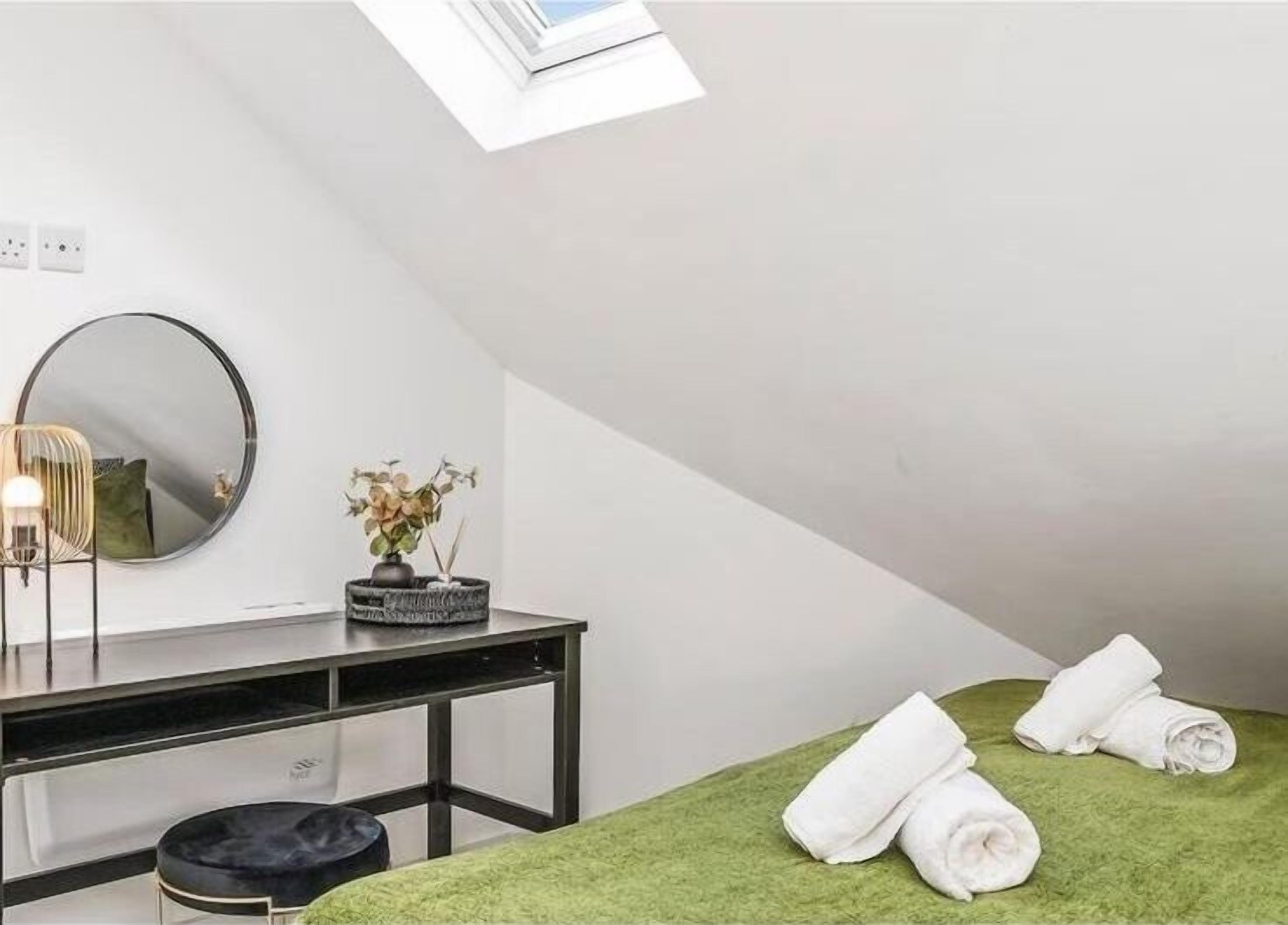 Une petite chambre moderne avec un puits de lumière, un miroir rond, une petite table ornée d’un vase de fleurs et un lit avec une literie verte et des serviettes enroulées.