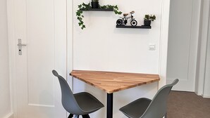 Una zona de menjador reduïda amb una taula de fusta i dues cadires, una porta blanca i dues estanteries amb plantes i una bicicleta.