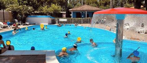 Una piscina con gente jugando waterpolo, rodeada de árboles y un edificio con tejado rojo.