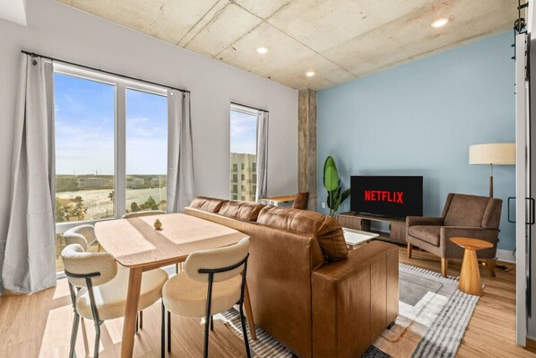 A modern living room with a sofa, dining table, chairs, a TV, and a view of the outdoors. - Stylish 1BR in Heart of Channelside (Tampa)