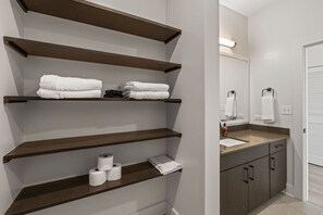 A bathroom with wooden shelves holding towels and toilet paper, a sink with a mirror, and a toilet. - Stylish 1BR in Heart of Channelside (Tampa)