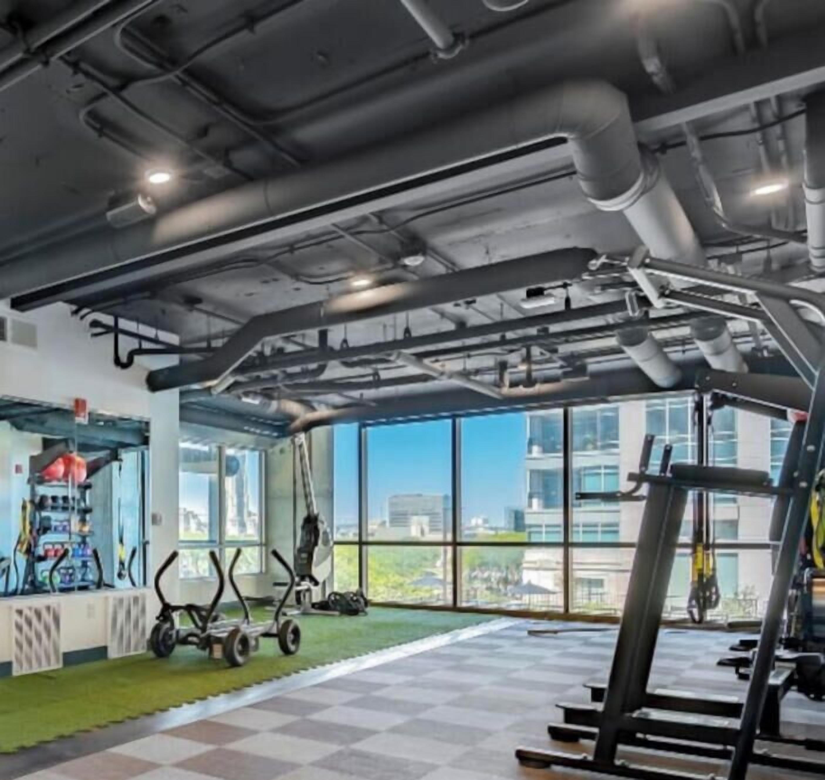 Ein Fitnessraum mit Trainingsgeräten, einem grünen Teppich und großen Fenstern mit Blick auf die Stadt.