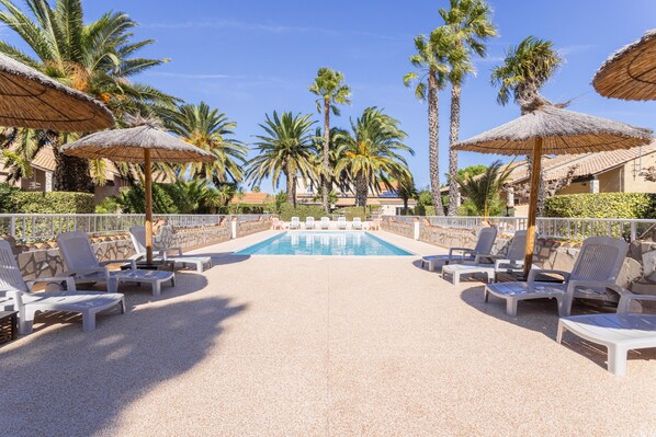 A pool area with lounge chairs and thatched umbrellas, surrounded by palm trees and a clear sky.