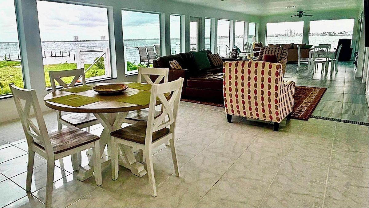 A spacious living area with a view of the water, a dining table with chairs, and a patterned armchair.
