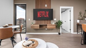 A modern living room with a mounted television displaying "NETFLIX," a dining area with a round table and chairs, and a balcony with a view. - A+ Spacious Luxury 2BR | Pool+Lounge+Parking (Indianapolis)