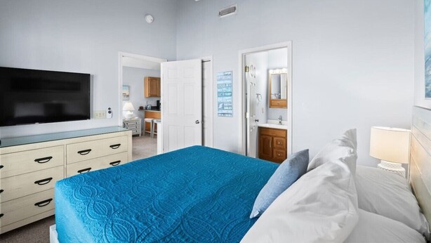 A bedroom with a bed, a television, a ceiling fan, a dresser, and a bathroom visible through an open door.
