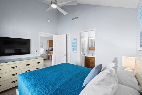 A bedroom with a bed, a television, a ceiling fan, a dresser, and a bathroom visible through an open door.