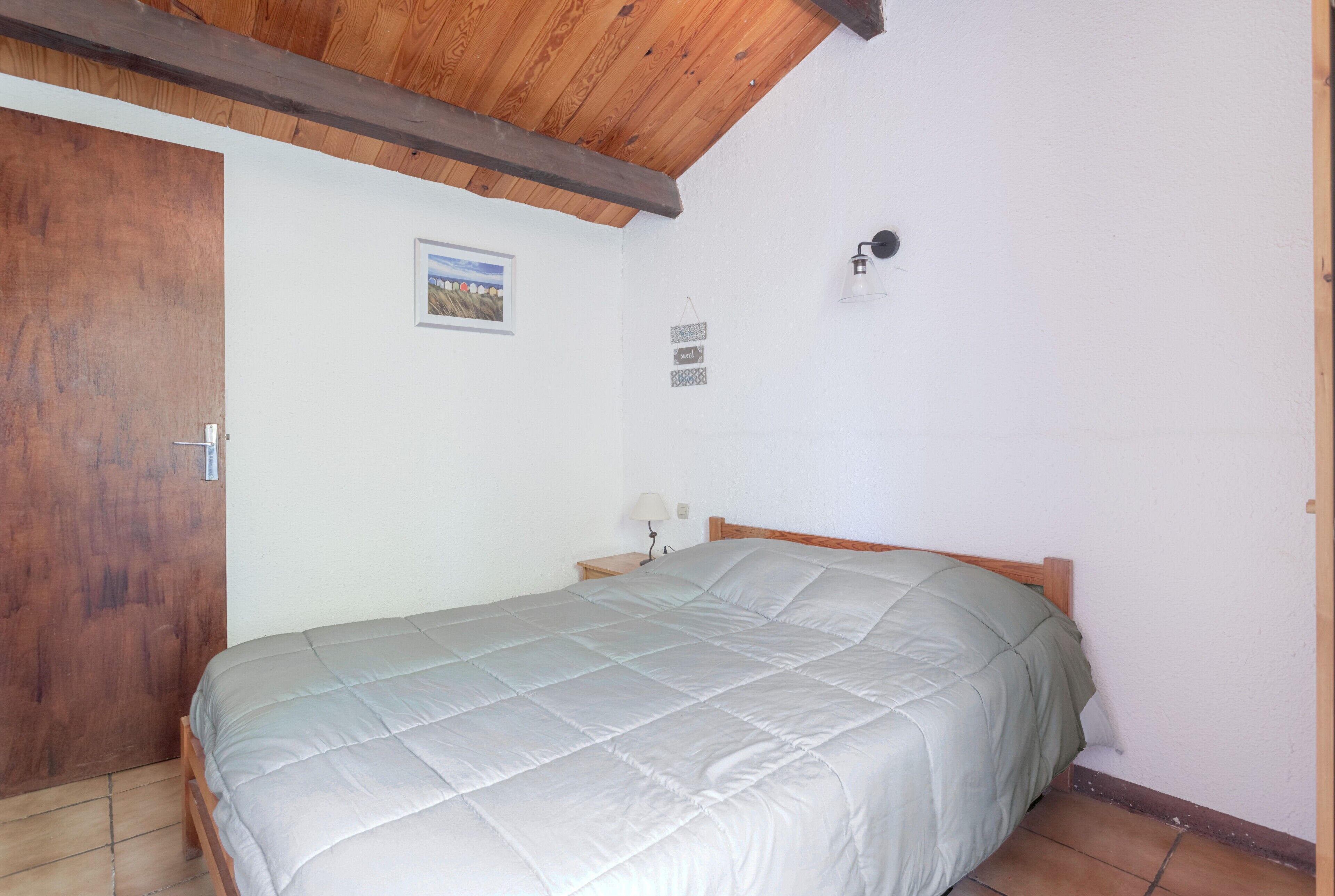 Une chambre à coucher avec un lit, des poutres en bois au plafond, une porte en bois, une table de chevet, une lampe fixée au mur et un tableau encadré.
