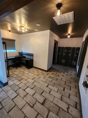 A room with a brick floor, a desk, a chair, a window with blinds, and a door leading to a locker area. - The Compound in downtown (Phoenix)