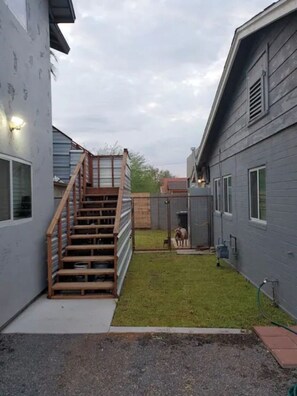 Ein Wohnhausgarten mit einer Holztreppe zu einer Terrasse, einem eingezäunten Bereich mit einem Hund und einem grauen Gebäude mit Fenstern.