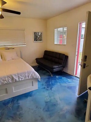 A bedroom with a bed, a black sofa, a ceiling fan, a window with blinds, and a door. - The Compound in downtown (Phoenix)