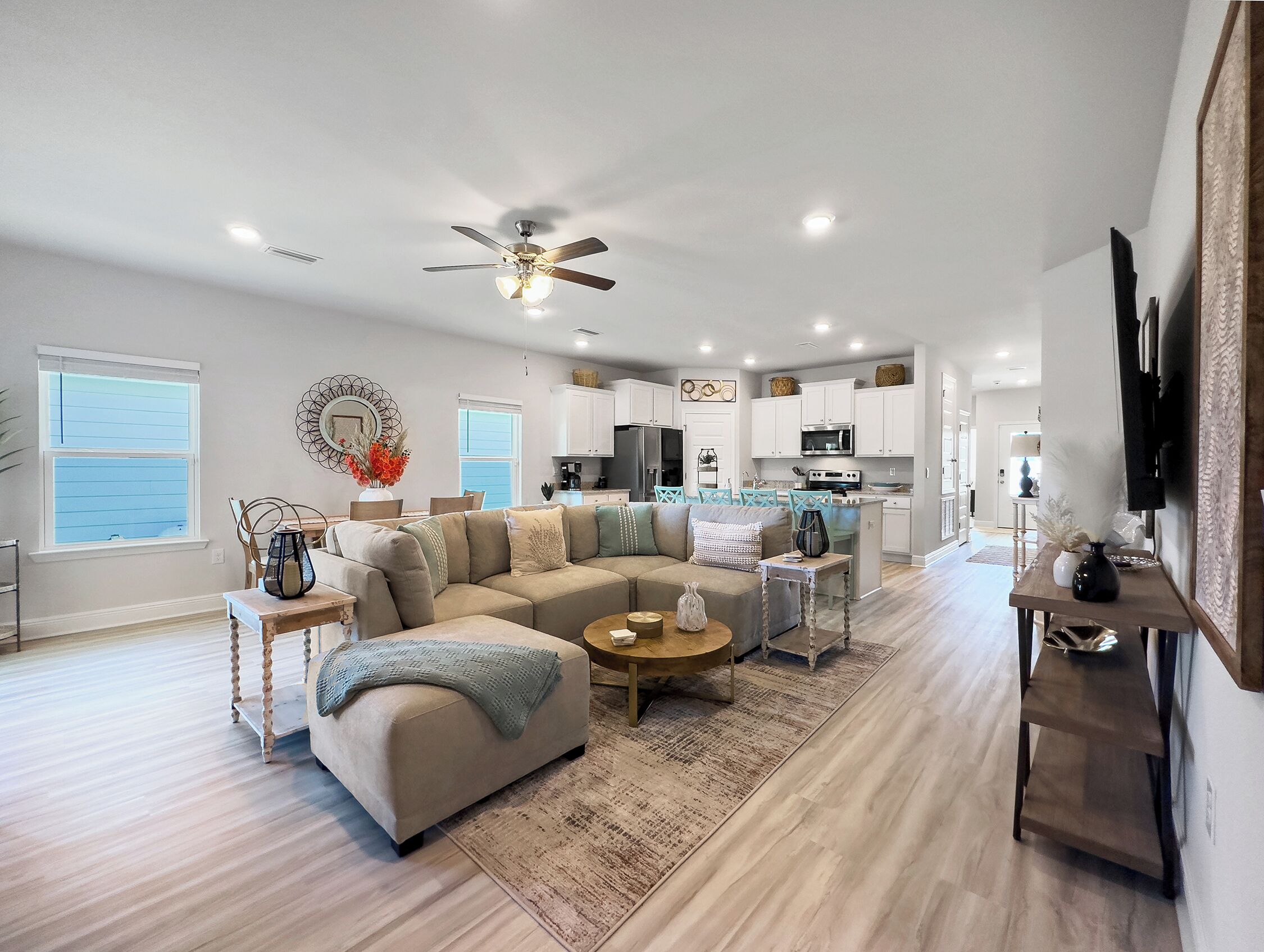 A spacious living room with a large sectional sofa, a coffee table, and a dining area with a small table and chairs. There is a kitchen in the background with white cabinets and a microwave.