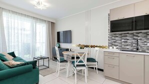 A modern living room with a green sofa, white dining chairs, and a kitchen area with a tiled backsplash and cabinets.