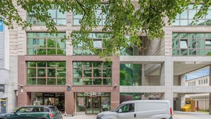 Un edificio moderno con una gran fachada de vidrio, un área de estacionamiento y una camioneta blanca estacionada al frente.