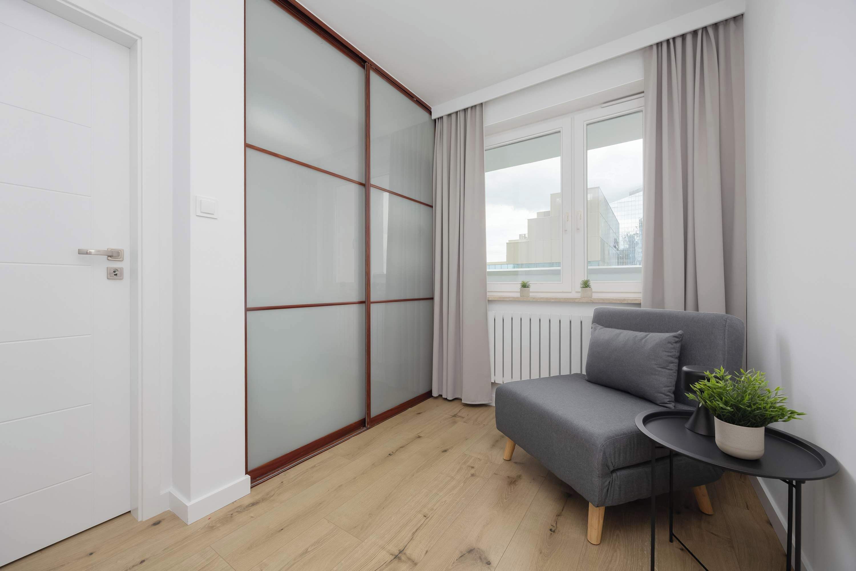 Una habitación moderna con puerta de vidrio corredera, un sofá gris, una mesita con una planta y vistas de la ciudad a través de la ventana.