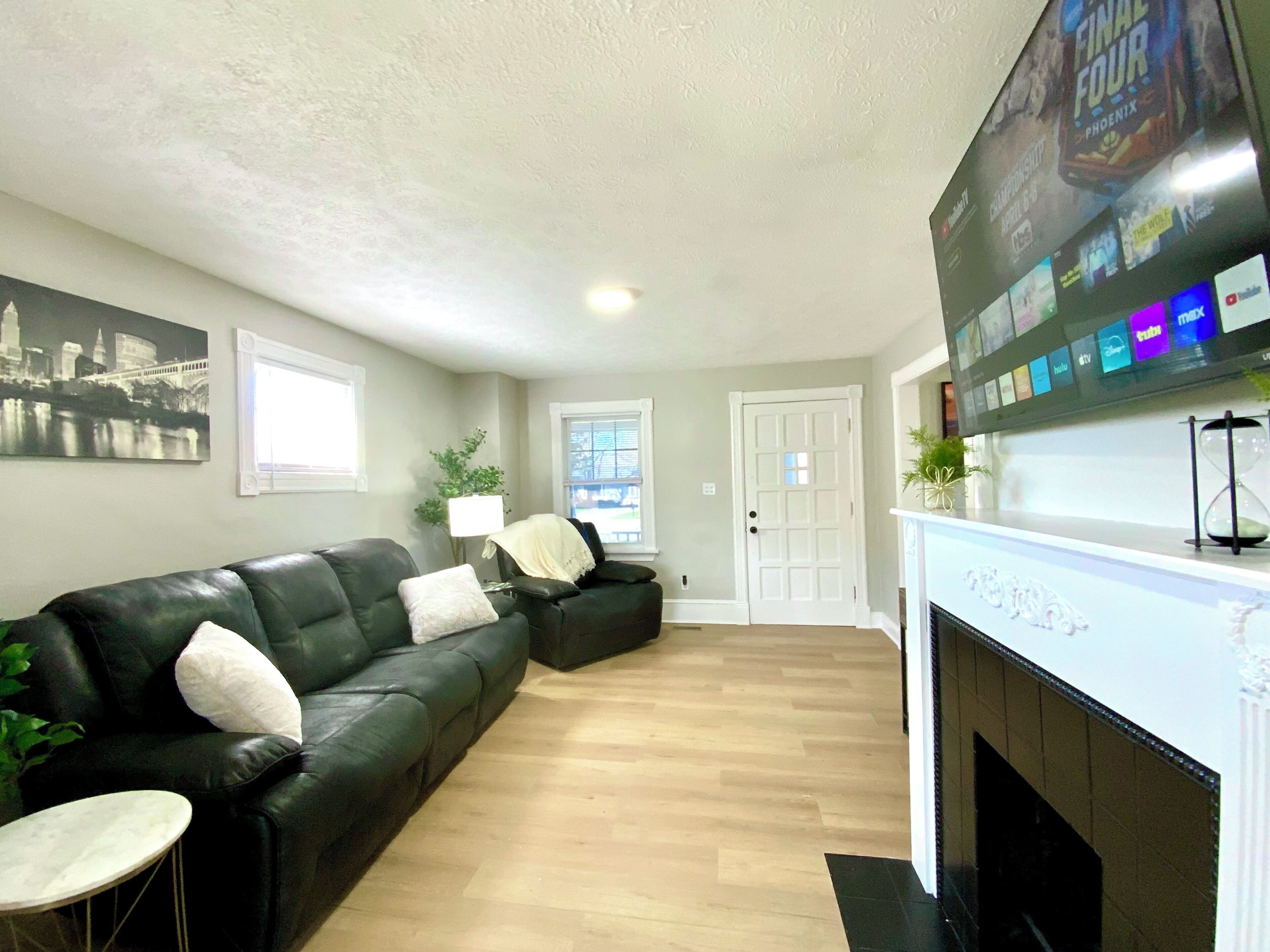 A living room with a black leather sofa, a small round table, a fireplace, and a wall-mounted TV displaying various channels.