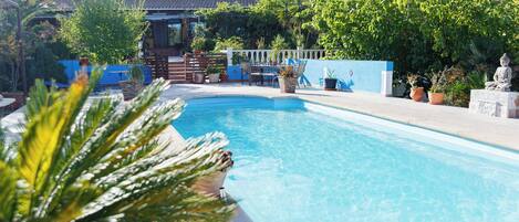 Une piscine avec un espace patio, un bâtiment doté d’un toit en tuiles et diverses plantes en pot.