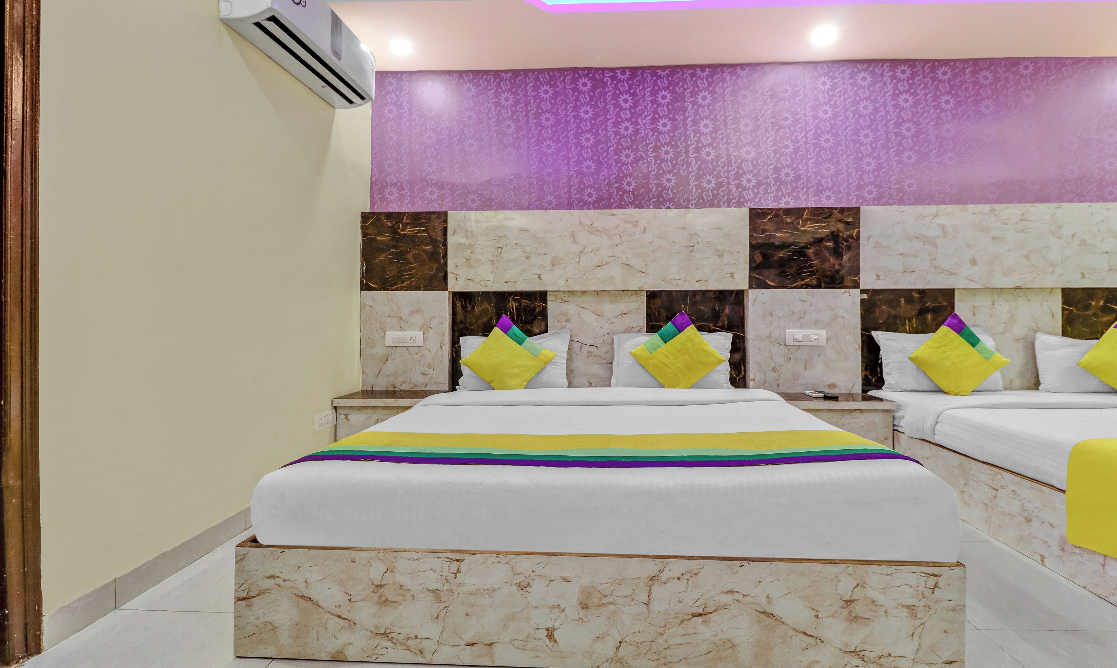 Une chambre d’hôtel avec un lit, deux tables de chevet, un climatiseur fixé au mur et un mur orné d’un motif violet.
