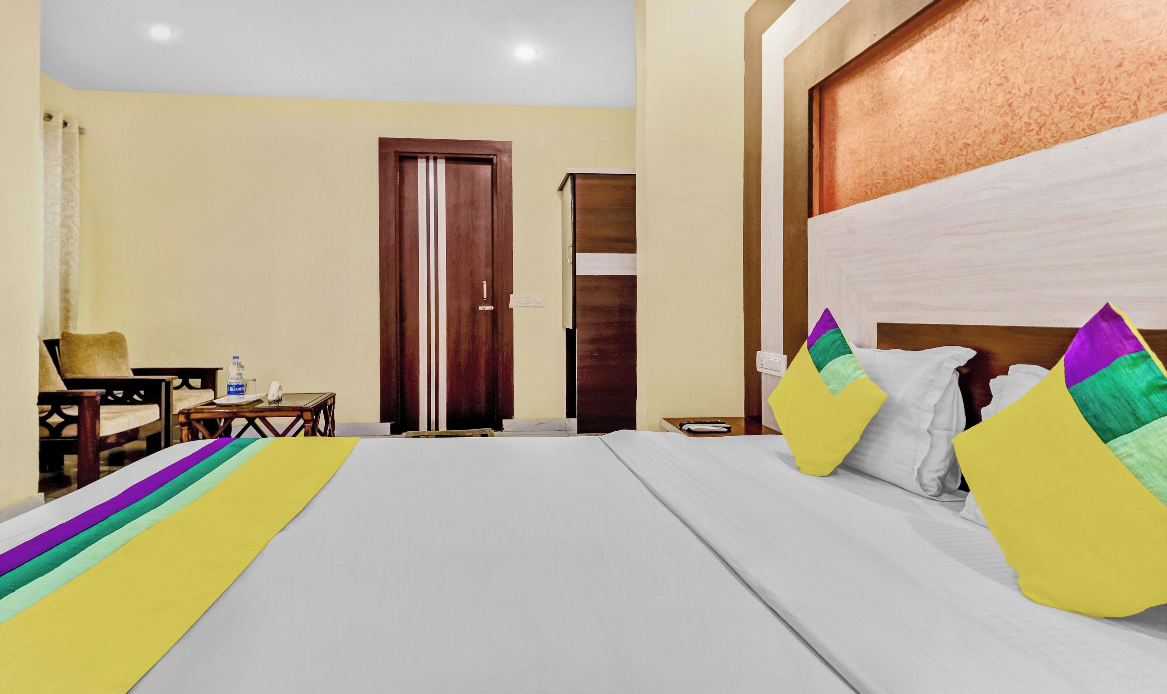 A hotel room with a large bed, a desk with a chair, a small table, and a door.