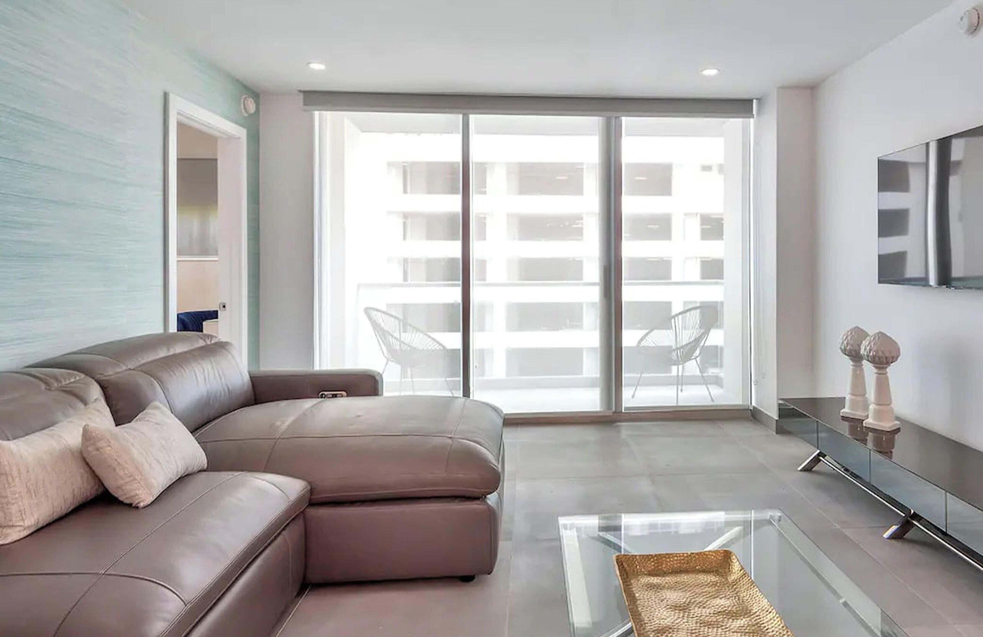 Una sala de estar moderna con un sofá de cuero marrón, una mesa de centro de vidrio y un gran ventanal corredizo que da a un balcón.