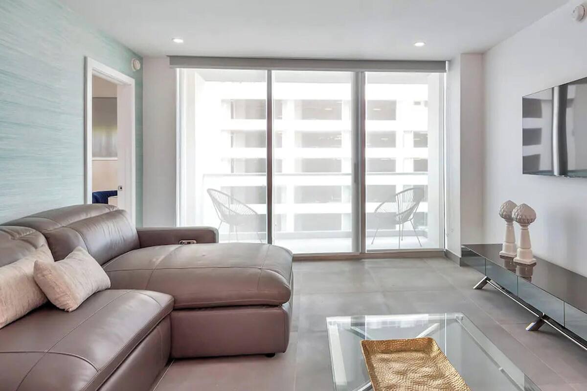A modern living room with a brown leather sofa, a glass coffee table, and a sliding glass door leading to a balcony.